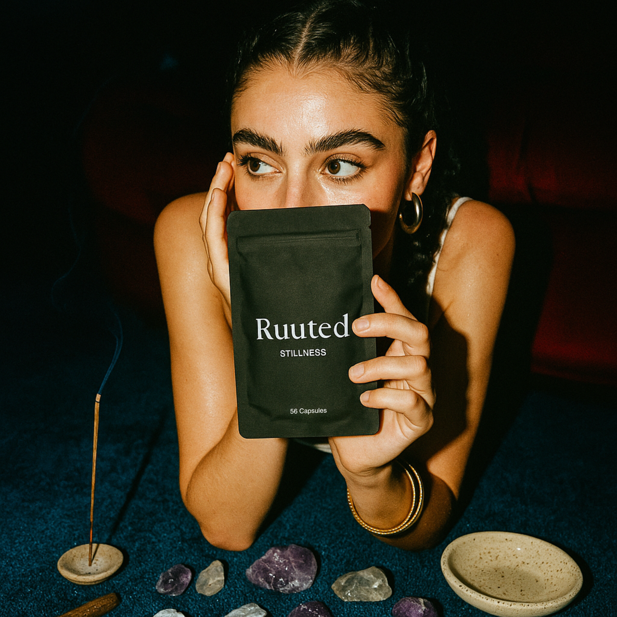 Person holding a black package labeled 'Ruuted Stillness' surrounded by stones and/incense.